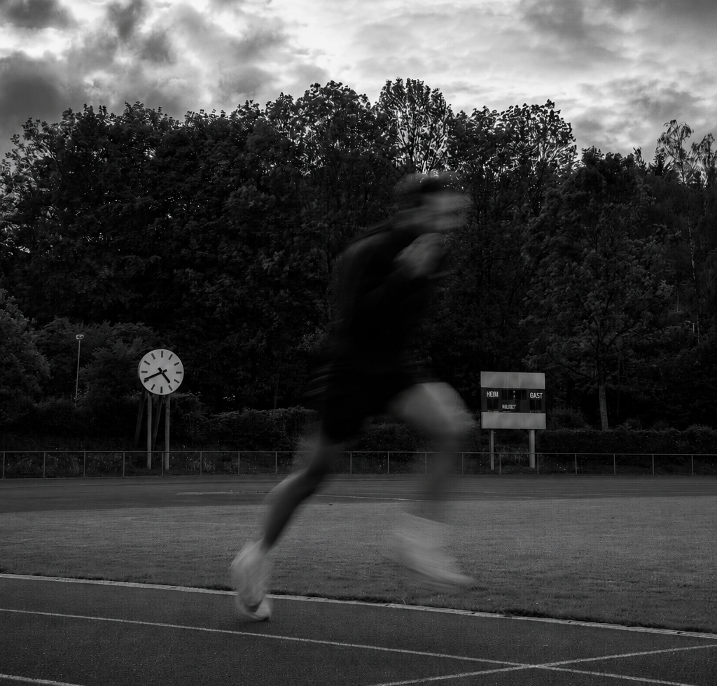 Schwarzweissaufnahme eines Sprinters in Bewegungsunschärfe auf der Tartanbahn neben einem Sportplatz mit Stadionuhr und Anzeigetafel.
