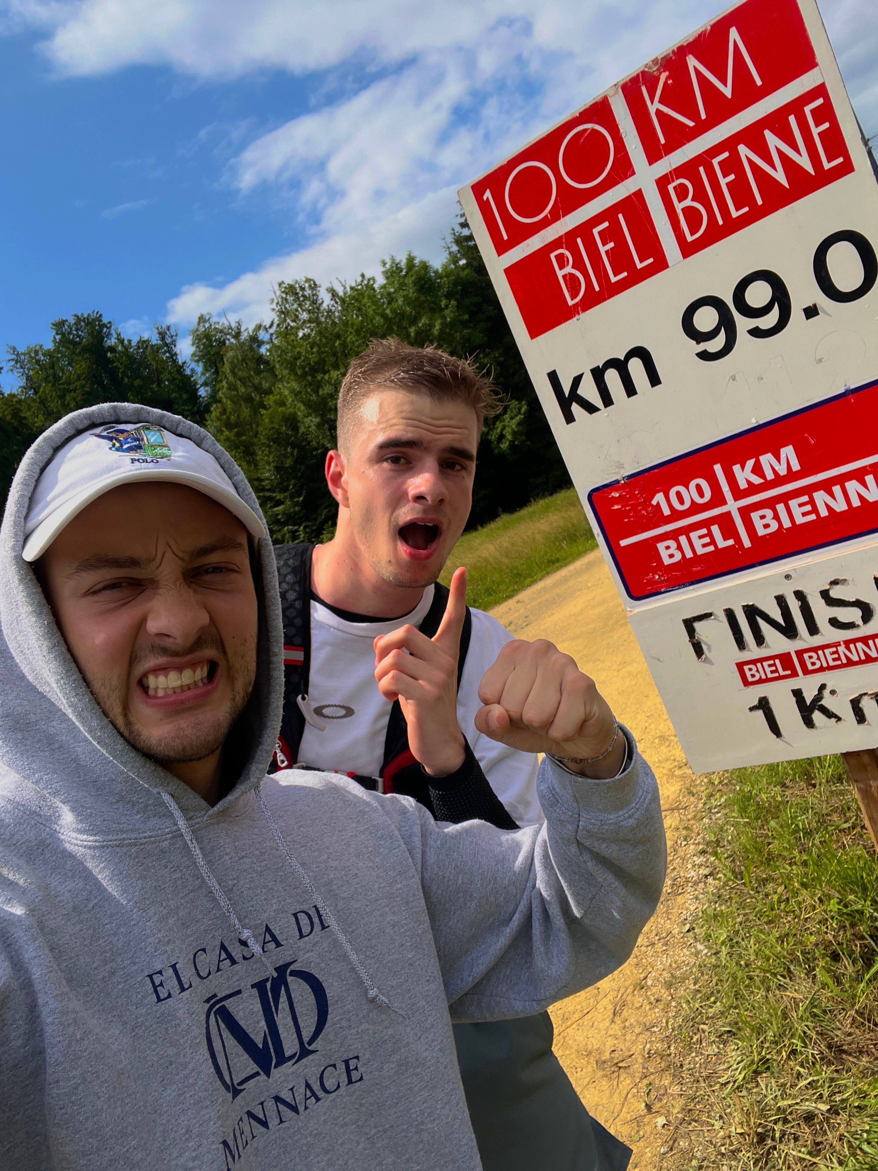 Zwei junge Athleten posieren am 100-km-Lauf Biel/Bienne kurz vor dem Ziel mit motivierten Gesten neben dem Streckenmarker bei sonnigem Wetter.