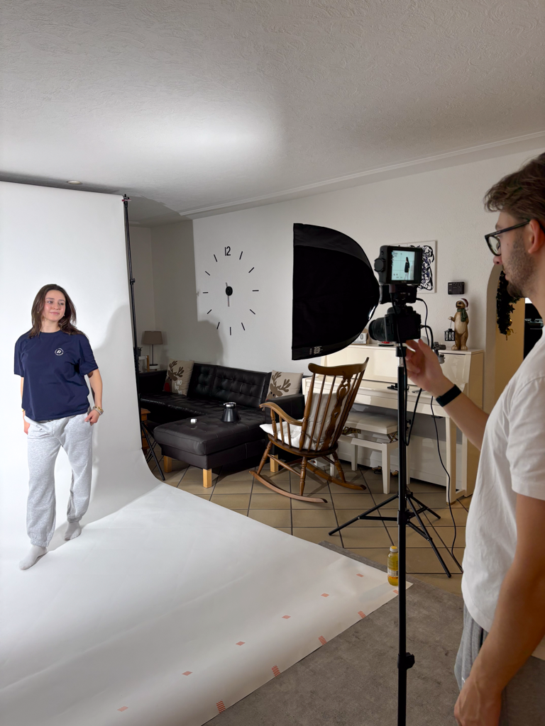Behind-the-Scenes-Aufnahme eines Athlix Fotoshootings mit Model im dunkelblauen T-Shirt vor weisser Studiokulisse und Fotograf mit Kamera.