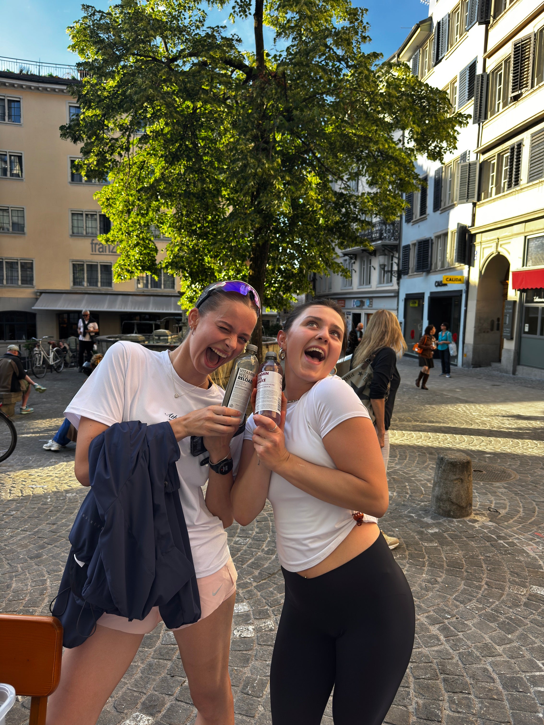 Zwei lachende Frauen in weissen Shirts feiern nach dem Lauf in der Altstadt und halten ihre Getränke in die Höhe.