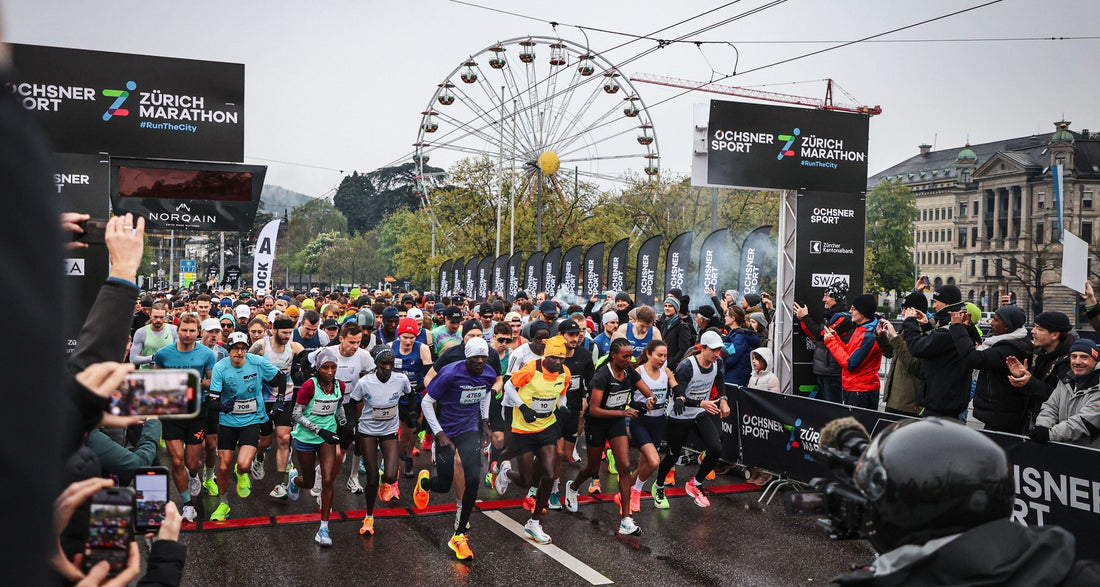 Massenstart des Zürich Marathons bei nasser Strasse: Hunderte Läuferinnen und Läufer starten zwischen Zuschauerreihen, grossen Event-Screens und einem Riesenrad im Hintergrund.