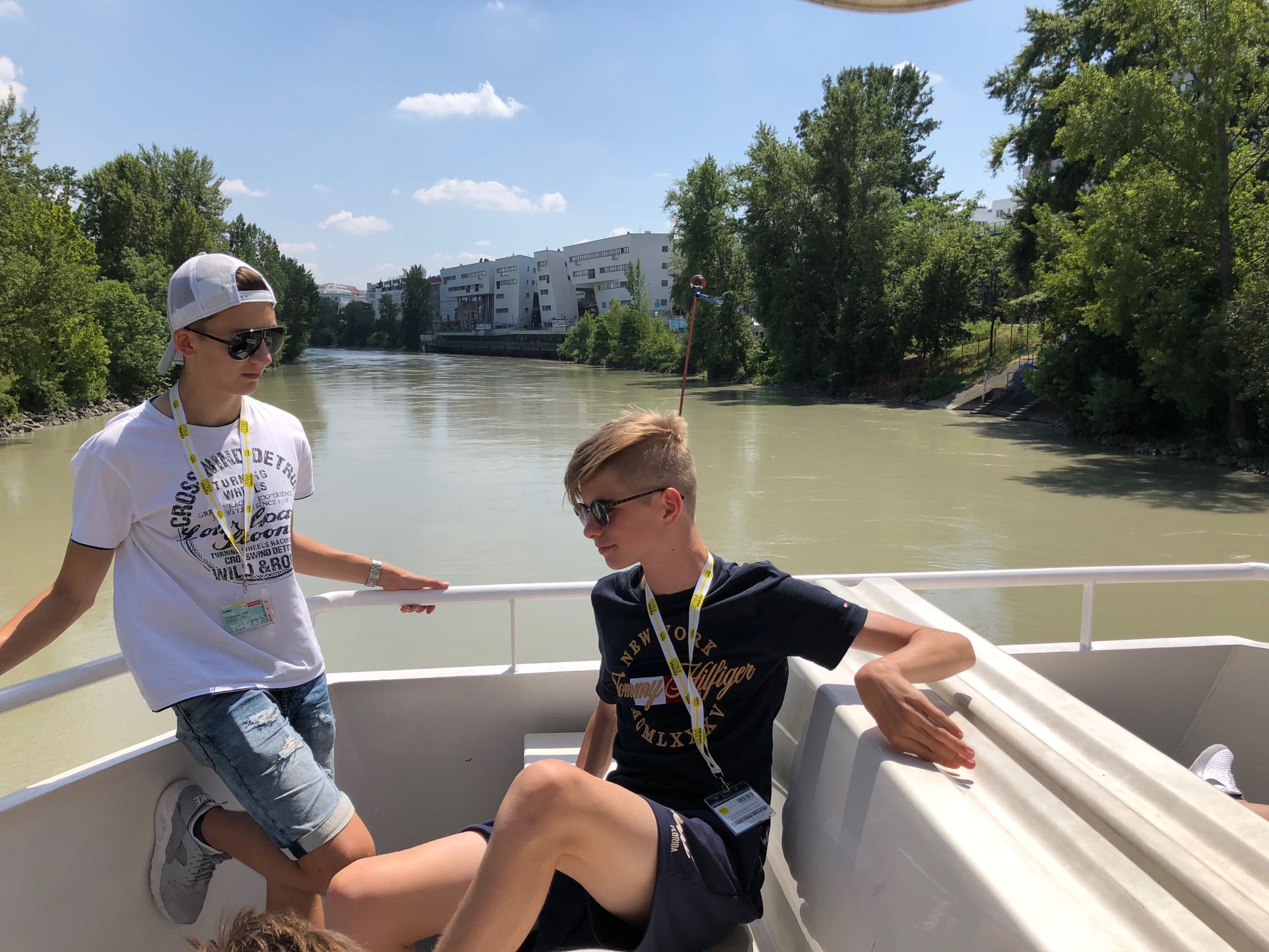 Zwei junge Männer geniessen einen sonnigen Sommertag auf einem Boot auf einem Fluss mit Grün und Stadtkulisse im Hintergrund.