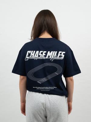Frau von hinten im dunkelblauen T-Shirt mit Chase-Miles-Print und Laufbahn-Grafik, dazu graue Jogginghose