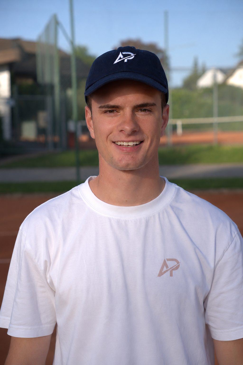 Junger Mann mit Athlix Cap und weissem T-Shirt mit AP Logo lächelt vor einem Sportplatz im Freien.