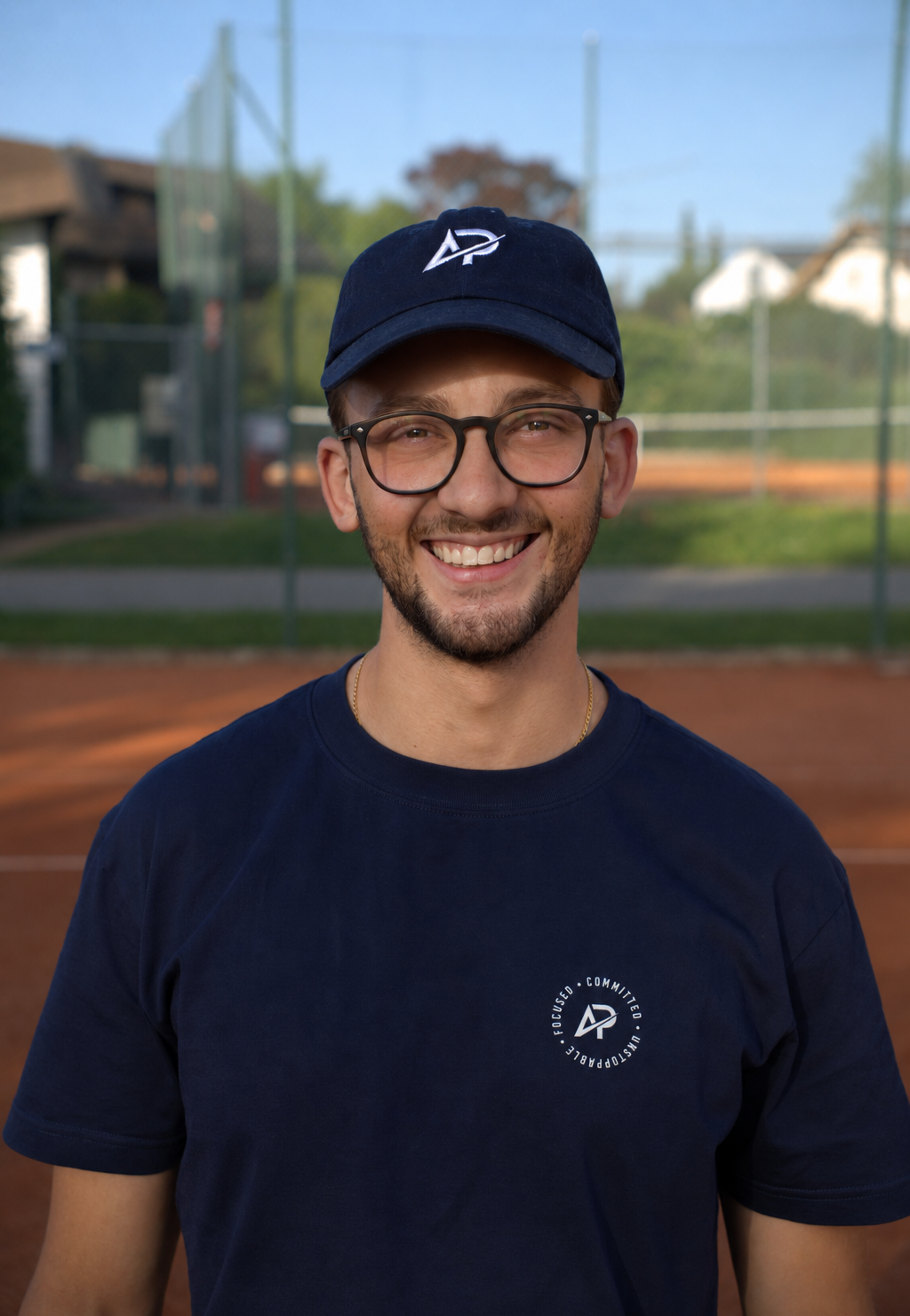 Junger Mann mit Athlix Cap und dunklem T-Shirt mit AP Logo lächelt auf einem Sportplatz im Freien.