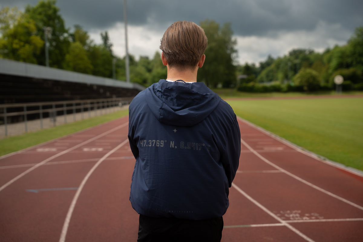 Läufer in dunkelblauer Trainingsjacke steht auf einer Tartanbahn und blickt konzentriert auf das leere Stadion.
