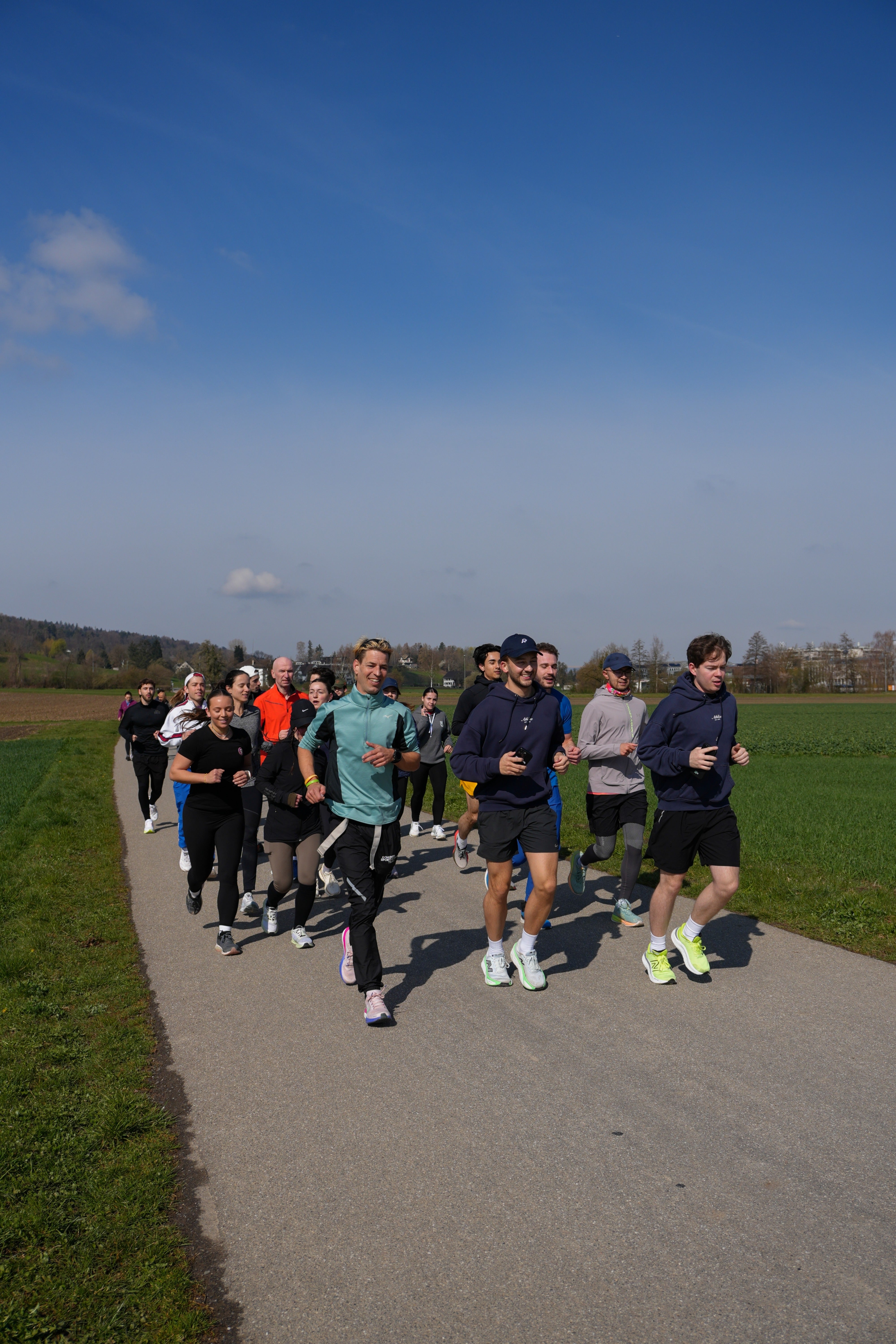 Grosse Laufgruppe in sportlicher Kleidung joggt gemeinsam auf einem Feldweg durch grüne Landschaft an einem sonnigen Tag mit blauem Himmel.